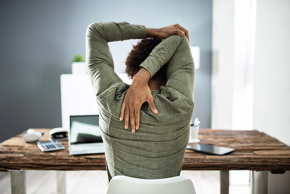 Three employees seated on chairs in an open office, arms folded overhead, following a LoDo yoga instructor seen from behind