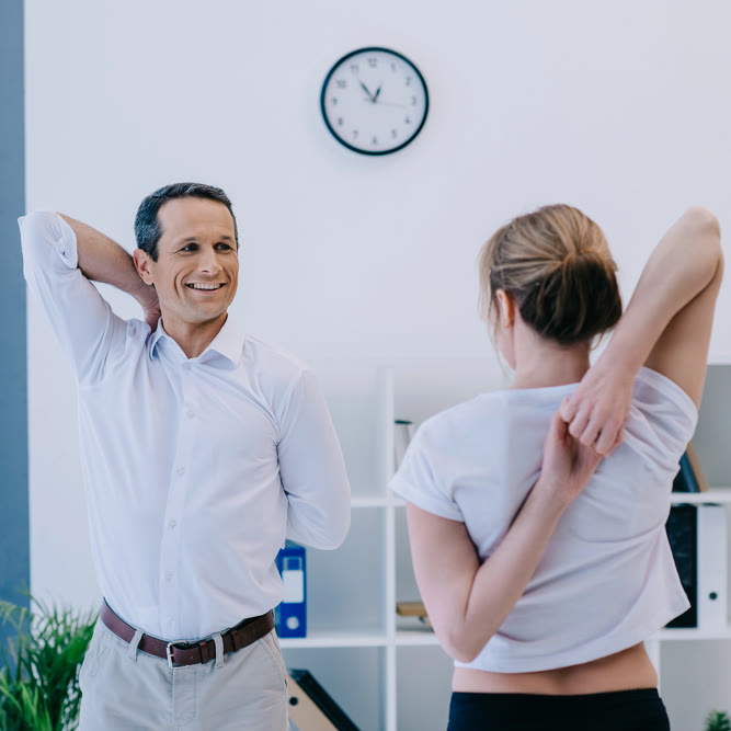 Two colleagues in work clothes stretching their arms behind their backs in a bright office